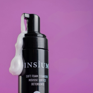 Black pump bottle with white foam clinging to its side, labeled softly as a cleanser, against a purple background.